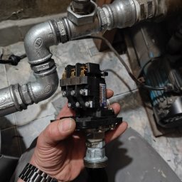 ELROI Robert Stankiewicz - Instalator trzyma w dłoni czarny stycznik elektryczny z widocznymi stykami, obok fragment instalacji wodnej z rurami ocynkowanymi i elementami złącznymi.