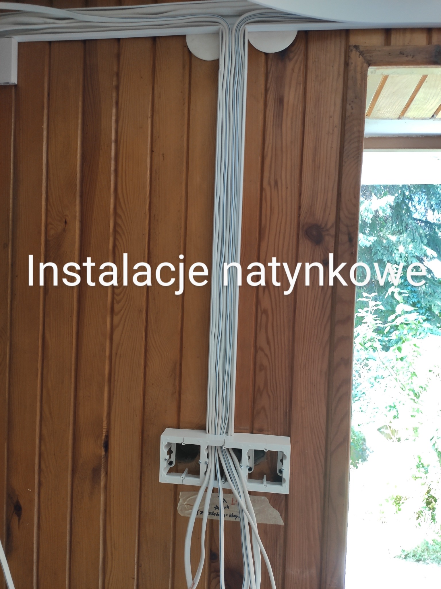Białe przewody elektryczne prowadzone natynkowo na drewnianej ścianie, z widoczną puszką instalacyjną i fragmentem okna z zielenią w tle.