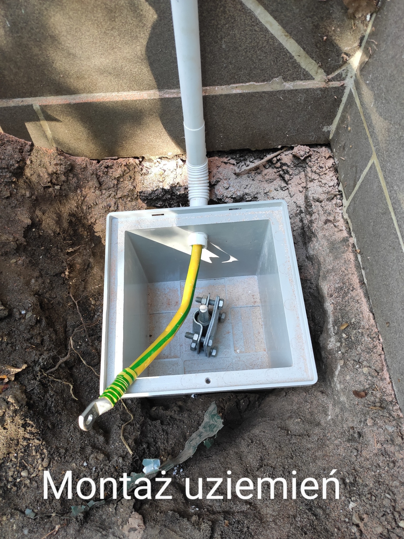 Uziemienie: Żółto-zielony przewód uziemiający z zaciskiem w szarej skrzynce, umieszczonej w wykopie przy fundamencie budynku.