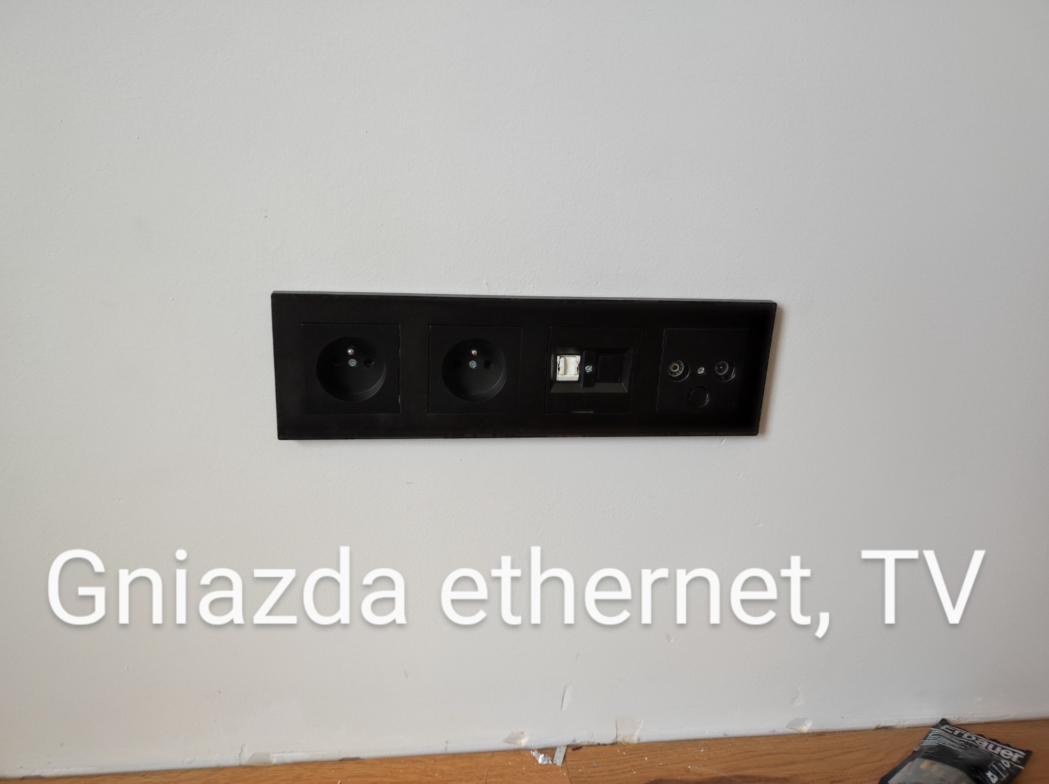 Czarny panel z gniazdami elektrycznymi, gniazdem sieciowym RJ45 i gniazdami antenowymi RTV zamontowany na białej ścianie nad drewnianą listwą przypodłogową.