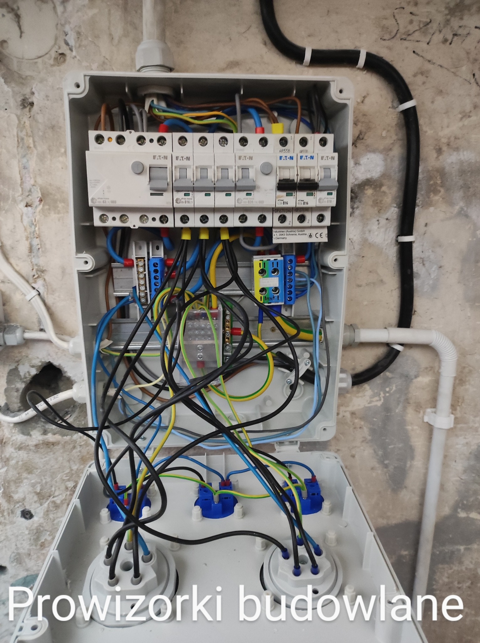 Otwarta skrzynka elektryczna z widocznymi bezpiecznikami EATON, przewodami w różnych kolorach i złączkami, zamontowana na surowej, otynkowanej ścianie, widoczne instalacje elektryczne tymczasowe.