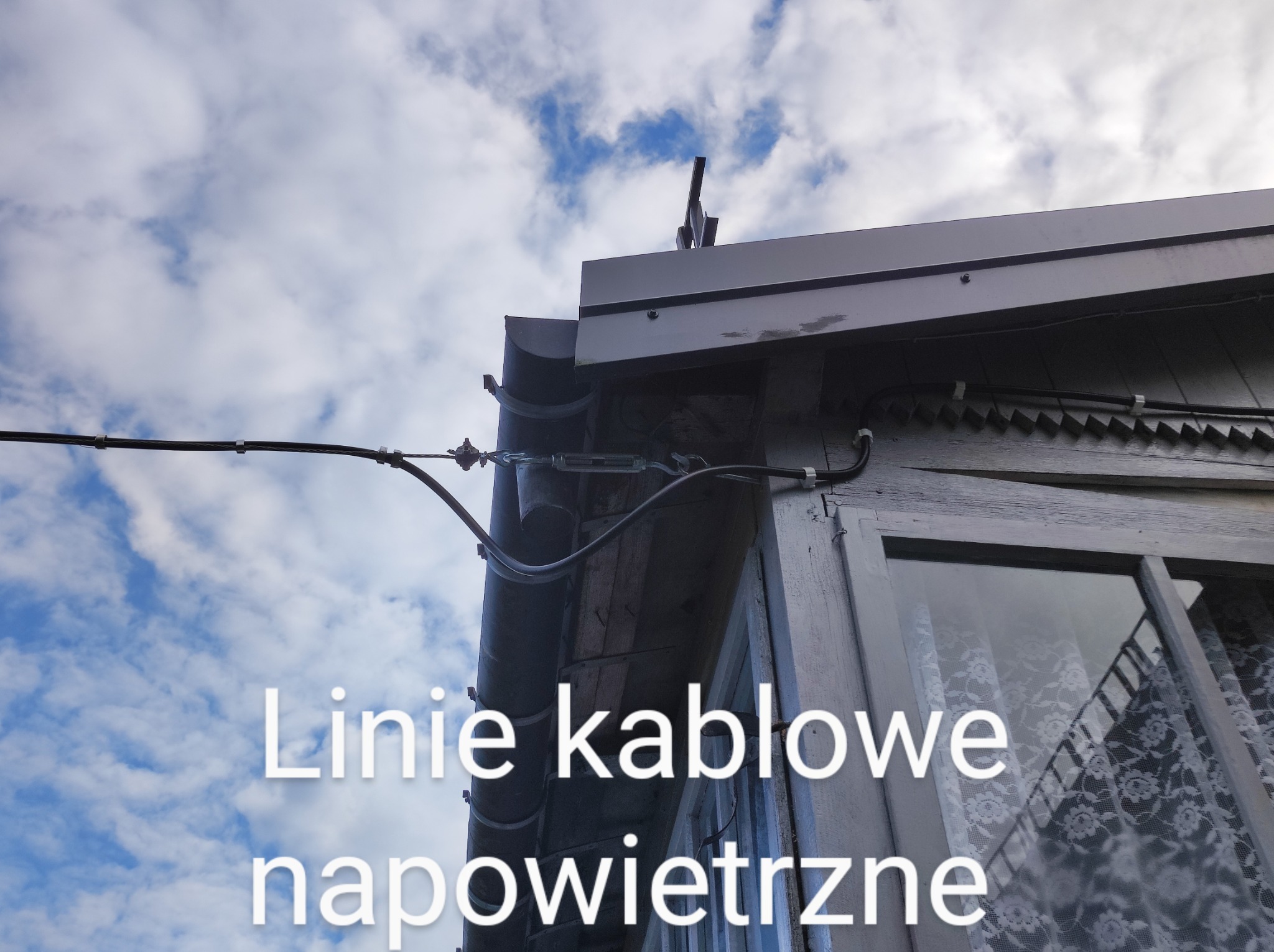 Napowietrzna linia kablowa przymocowana do drewnianego budynku z widocznym oknem i metalową rynną na tle błękitnego nieba z chmurami.