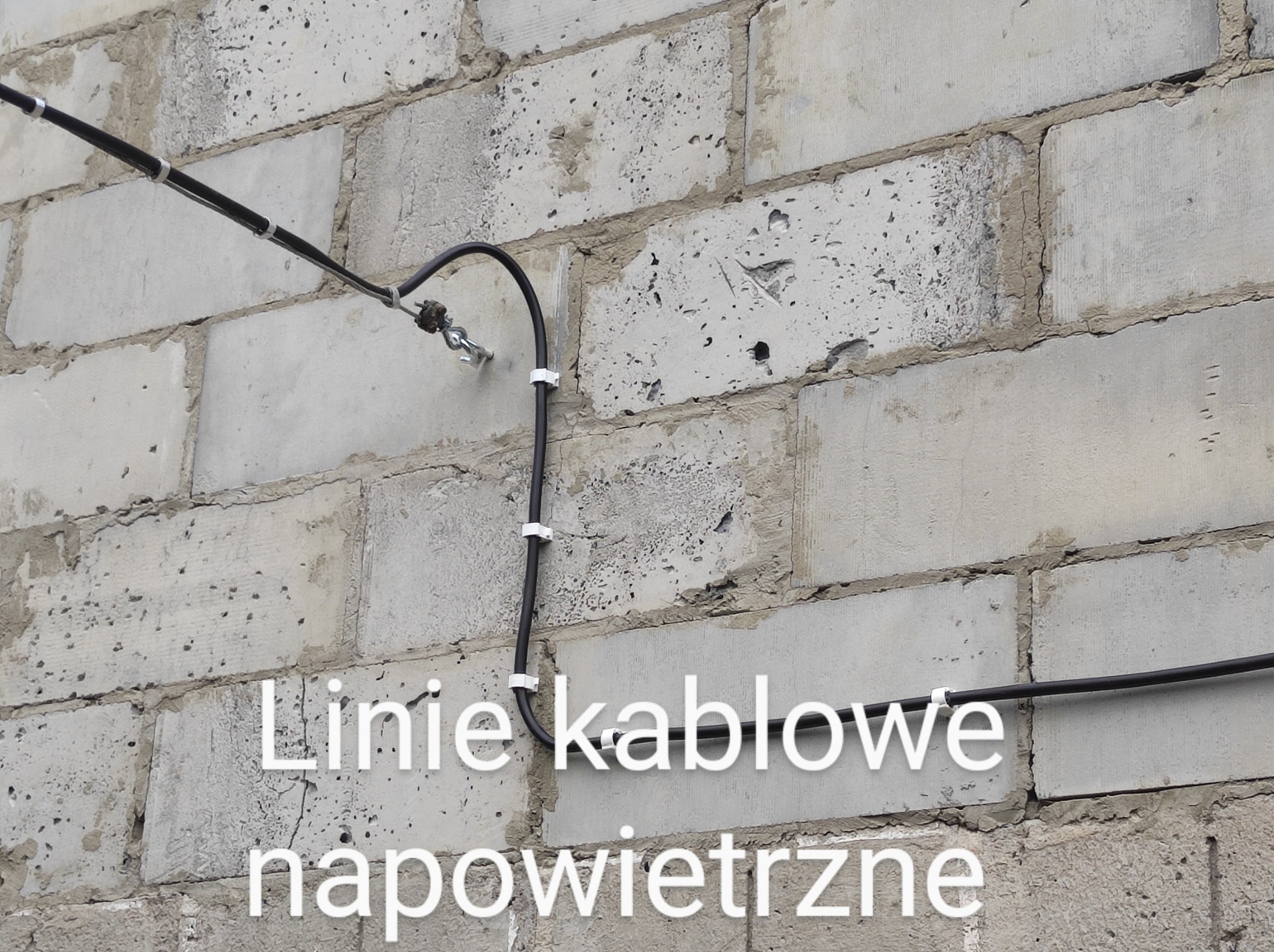 Czarny kabel elektryczny przymocowany do ściany z bloczków betonowych za pomocą białych uchwytów, widoczne złącze śrubowe.