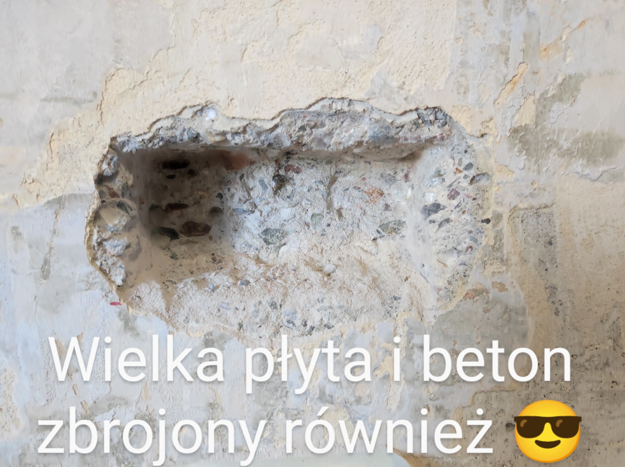 Odsłonięty fragment betonowej ściany z widocznym zbrojeniem i kruszywem, otoczony resztkami starego tynku.