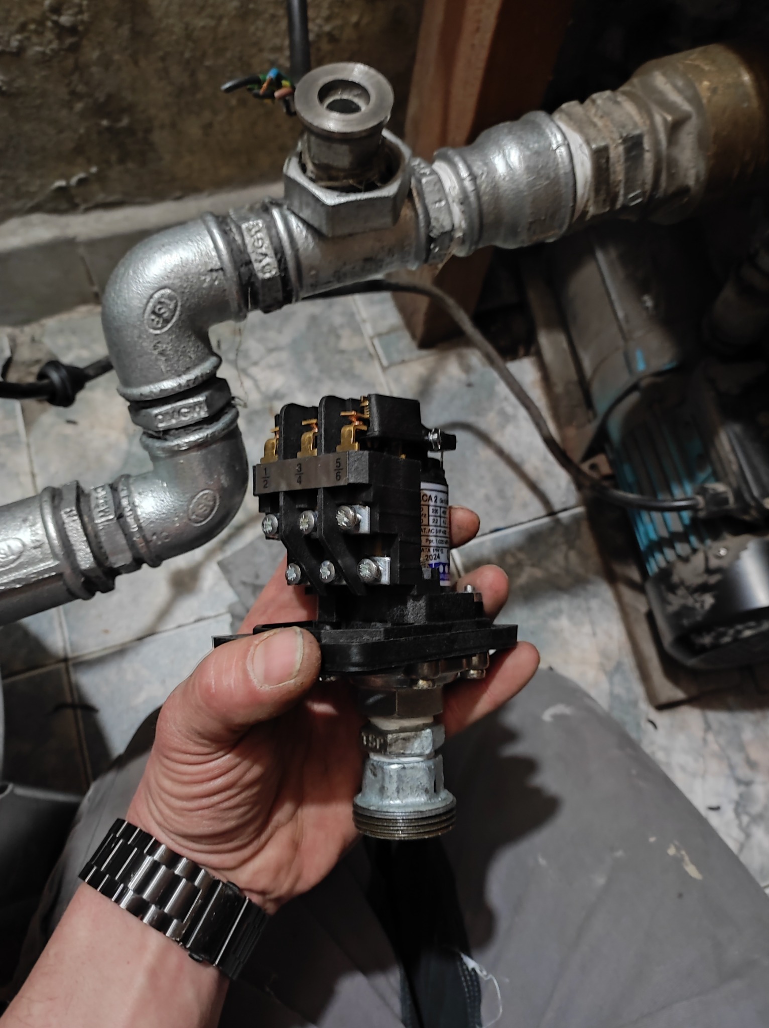 Instalator trzyma w dłoni czarny stycznik elektryczny z widocznymi stykami, obok fragment instalacji wodnej z rurami ocynkowanymi i elementami złącznymi.