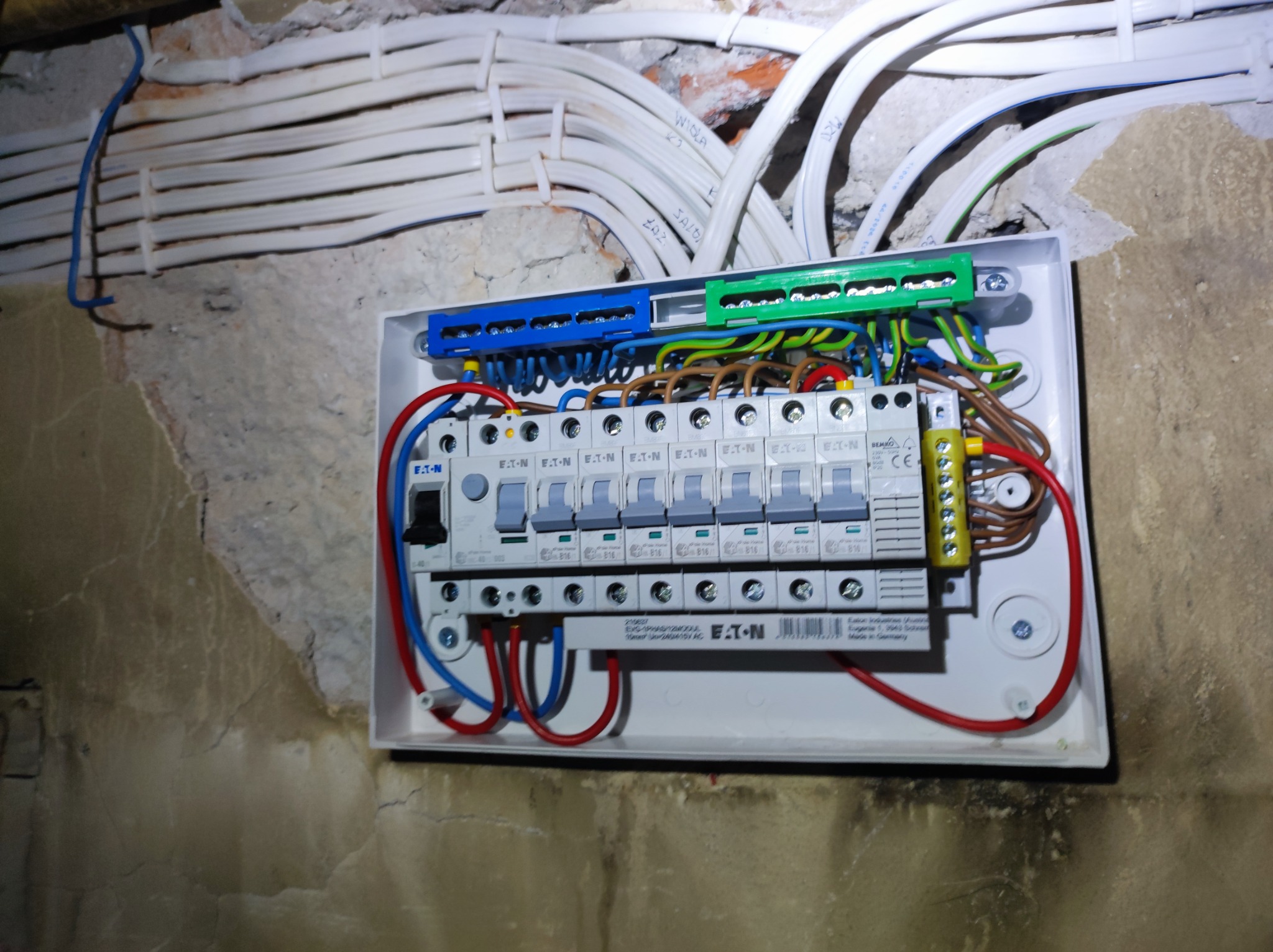 Rozdzielnica elektryczna firmy Eaton zamontowana na surowej ścianie z wyprowadzonymi przewodami w peszlach, widoczne listwy zaciskowe w kolorach niebieskim i zielonym.