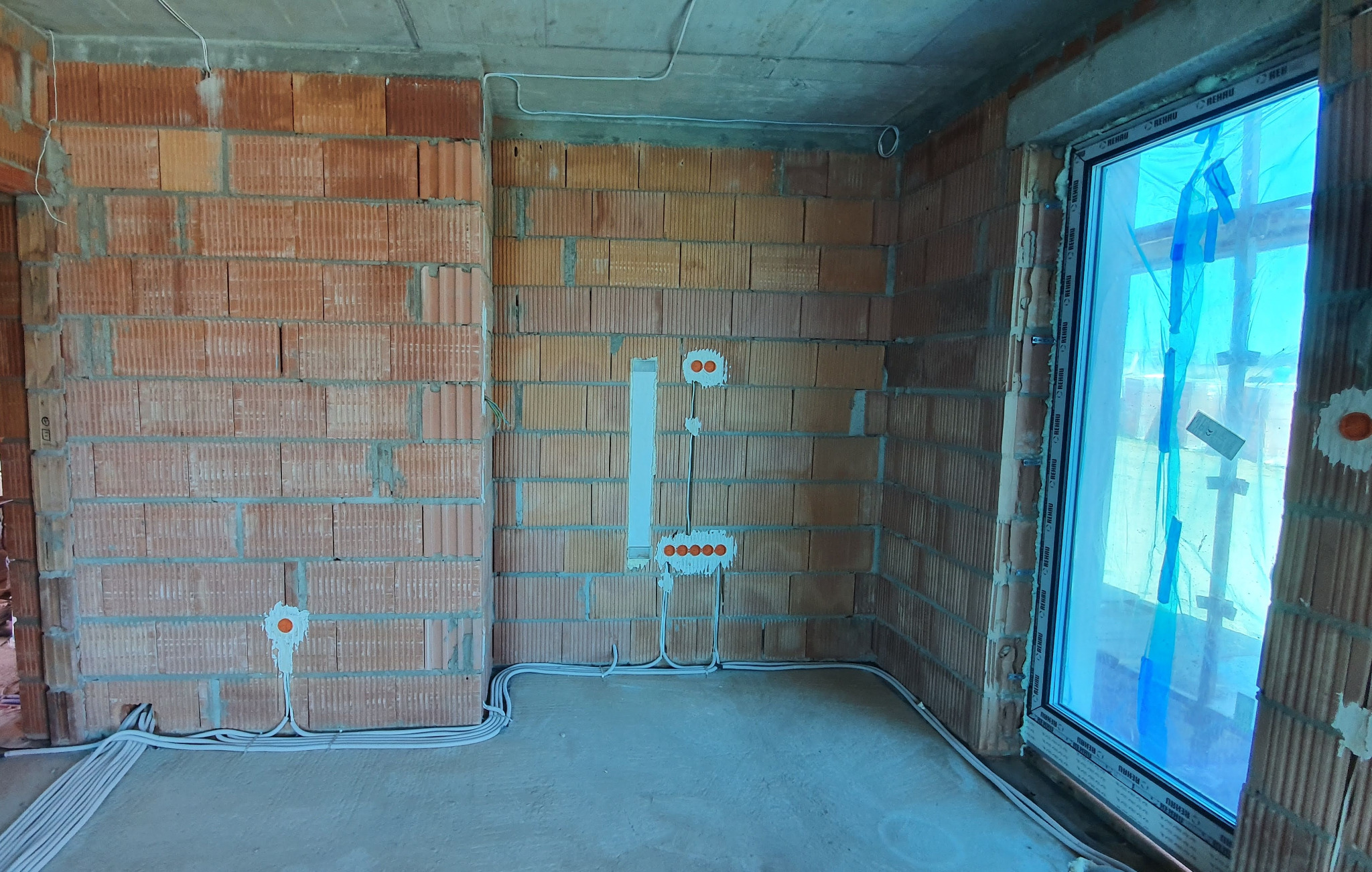 Surowy stan wnętrza z ceglanymi ścianami, widoczne przygotowane punkty elektryczne i rozprowadzone kable wzdłuż podłogi, okno zabezpieczone folią.