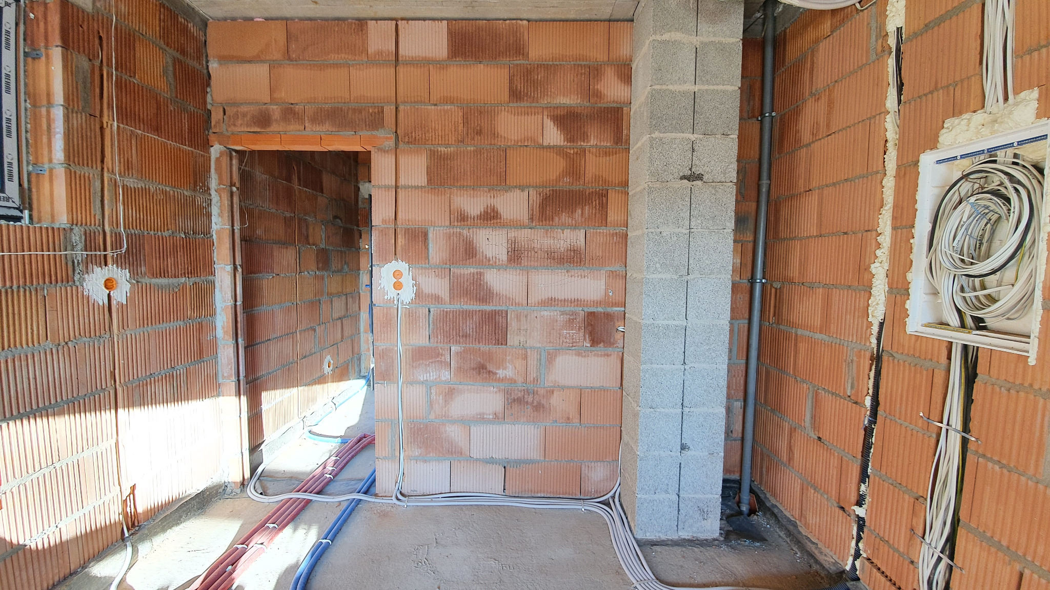 Wnętrze w surowym stanie z widocznymi instalacjami elektrycznymi: kable prowadzone po podłodze i w ścianach z cegły dziurawki, puszka elektryczna z przewodami oraz fragment szarej kolumny betonowej...