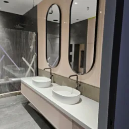 Nowoczesna łazienka w trakcie wykończenia: dwie owalne umywalki nablatowe, czarne baterie, podwieszana szafka z ryflowanym frontem, dwa owalne lustra w czarnych ramach, ściany wyłożone pionową...