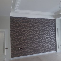 MERIDIAN EU SPÓŁKA Z OGRANICZONĄ ODPOWIEDZIALNOŚCIĄ - Pokój po remoncie: ściana pokryta wzorzystą tapetą w odcieniach brązu i beżu, białe listwy przypodłogowe i sufitowe, białe drzwi z ozdobnymi panelami, gładka podłoga, widoczne gniazdka elektryczne.