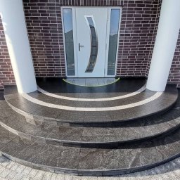 MERIDIAN EU SPÓŁKA Z OGRANICZONĄ ODPOWIEDZIALNOŚCIĄ - Eleganckie, granitowe schody wejściowe z białymi akcentami, prowadzące do nowoczesnych, białych drzwi z przeszkleniami i brązowej ceglanej elewacji.