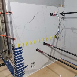 MERIDIAN EU SPÓŁKA Z OGRANICZONĄ ODPOWIEDZIALNOŚCIĄ - Montaż wielkoformatowych płyt imitujących marmur w łazience, użycie przyssawek i rozpórek do utrzymania płyty, widoczne poziomowanie z użyciem klipsów i pasków niebieskiej taśmy malarskiej...