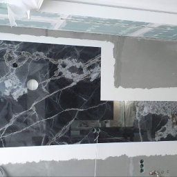 MERIDIAN EU SPÓŁKA Z OGRANICZONĄ ODPOWIEDZIALNOŚCIĄ - Montaż paneli z czarnego marmuru z białymi żyłami na ścianie w trakcie prac wykończeniowych, widoczne wystające kable elektryczne i surowy tynk.