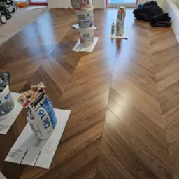 Układanie parkietu w jodełkę z widocznymi materiałami budowlanymi: worki z klejem i wiaderka z masą szpachlową, kartony ochronne na podłodze, okna w tle.