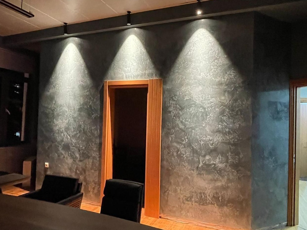 Szara ściana z efektem dekoracyjnym, oświetlona trzema punktowymi lampami sufitowymi, z widocznym fragmentem drewnianej ramy drzwiowej i elementami wyposażenia wnętrza.