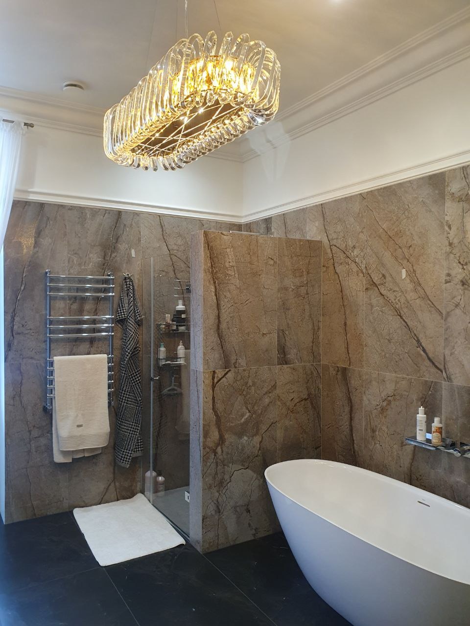 Nowoczesna łazienka z marmurowymi płytkami, wolnostojącą białą wanną i designerskim, kryształowym żyrandolem w kształcie prostokąta.