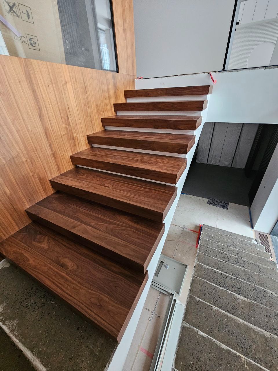 Nowoczesne schody z drewnianymi stopniami o ciepłym, orzechowym odcieniu, kontrastujące z białymi podstopnicami, prowadzące w dół; widoczna surowa betonowa konstrukcja schodów i drewniana okładzina...