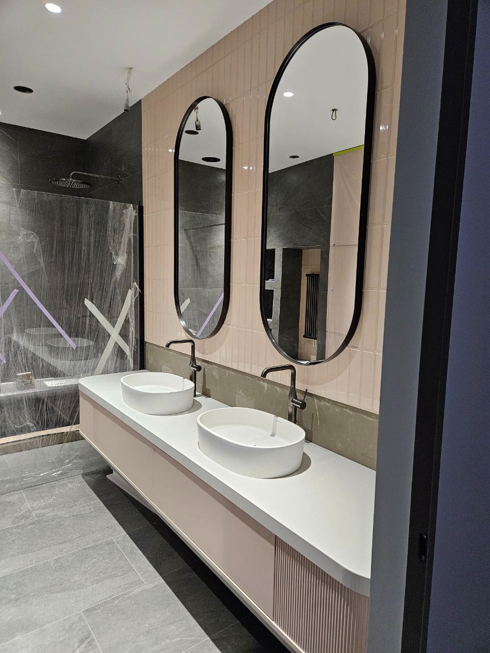 Nowoczesna łazienka w trakcie wykończenia: dwie owalne umywalki nablatowe, czarne baterie, podwieszana szafka z ryflowanym frontem, dwa owalne lustra w czarnych ramach, ściany wyłożone pionową...