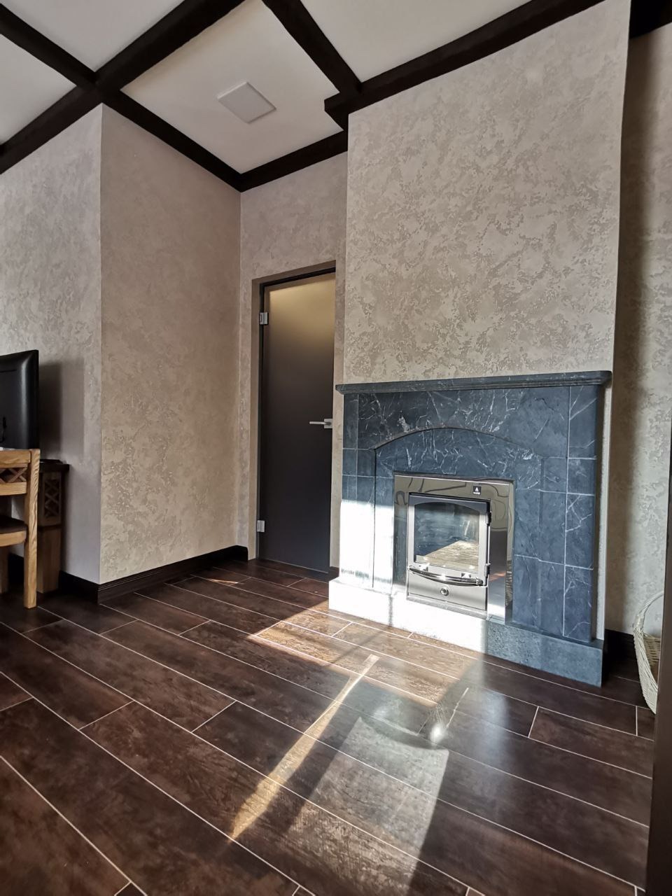 Nowoczesny salon z kominkiem obudowanym ciemnym marmurem, dekoracyjna faktura ścian w odcieniach beżu, ciemnobrązowa podłoga z paneli imitujących drewno, sufit z ciemnymi belkami.