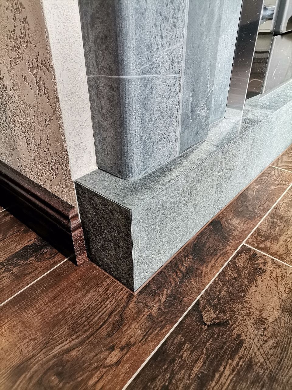 Detale wykończenia łazienki: ciemnobrązowe płytki podłogowe imitujące drewno, szara, kamienna obudowa wanny lub brodzika oraz dekoracyjna ściana z fakturą.