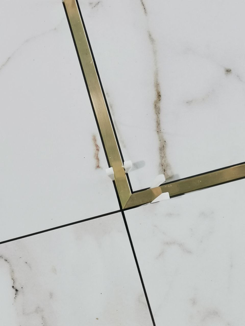 Białe płytki imitujące marmur z metalowymi profilami narożnymi w kolorze złotym, zabezpieczone plastikowymi krzyżykami dystansowymi.