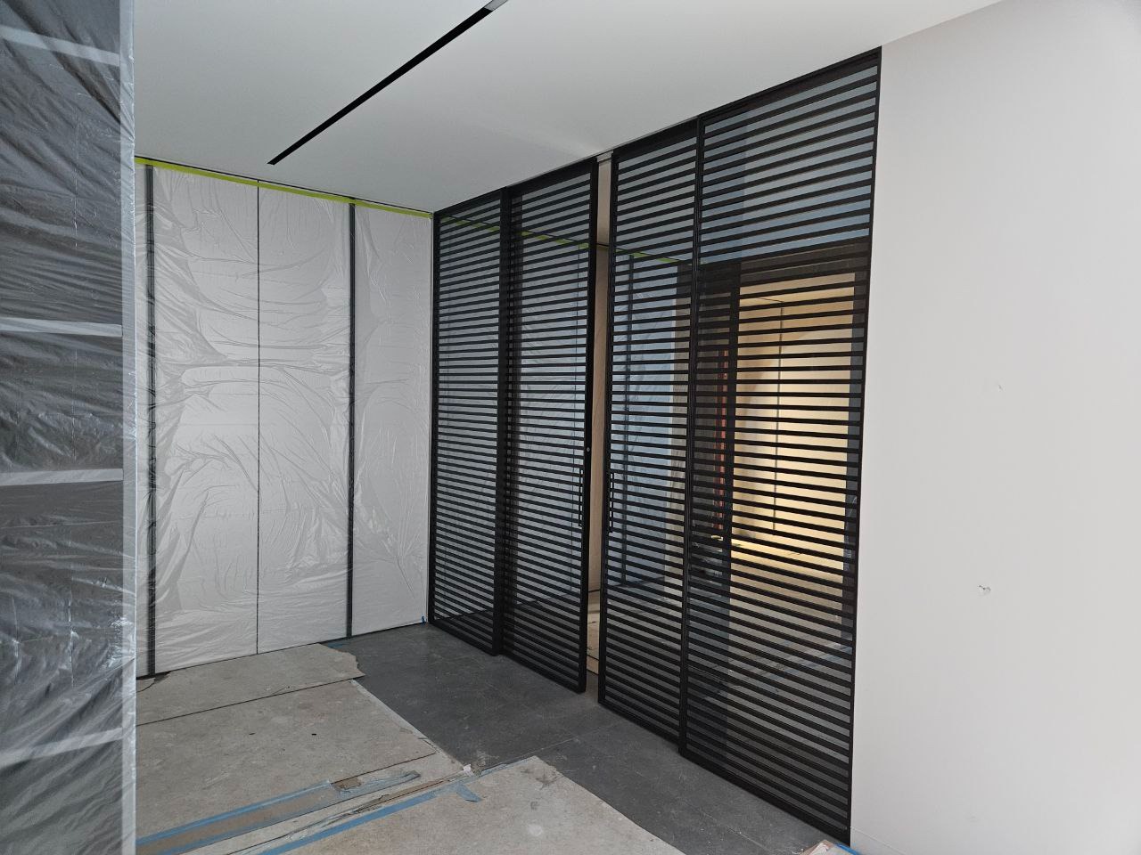 Wnętrze w trakcie prac wykończeniowych: ściany zabezpieczone folią malarską, widoczne czarne, poziome żaluzje oddzielające przestrzeń.
