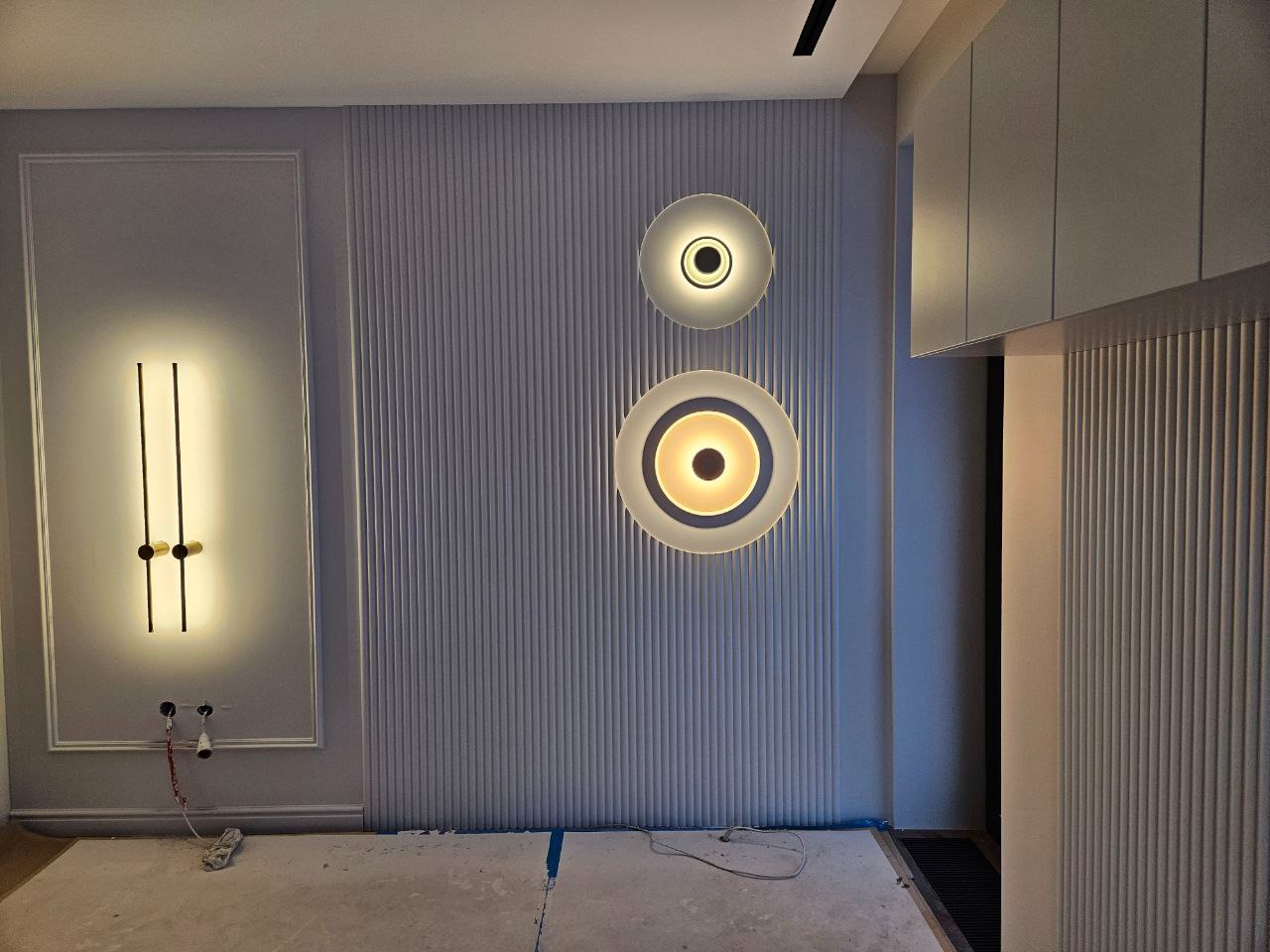 Nowoczesne oświetlenie ścienne: dwa okrągłe panele LED oraz pionowa lampa z czarnymi prętami na tle szarej ściany z pionowymi żłobieniami, widoczne niedokończone prace wykończeniowe.