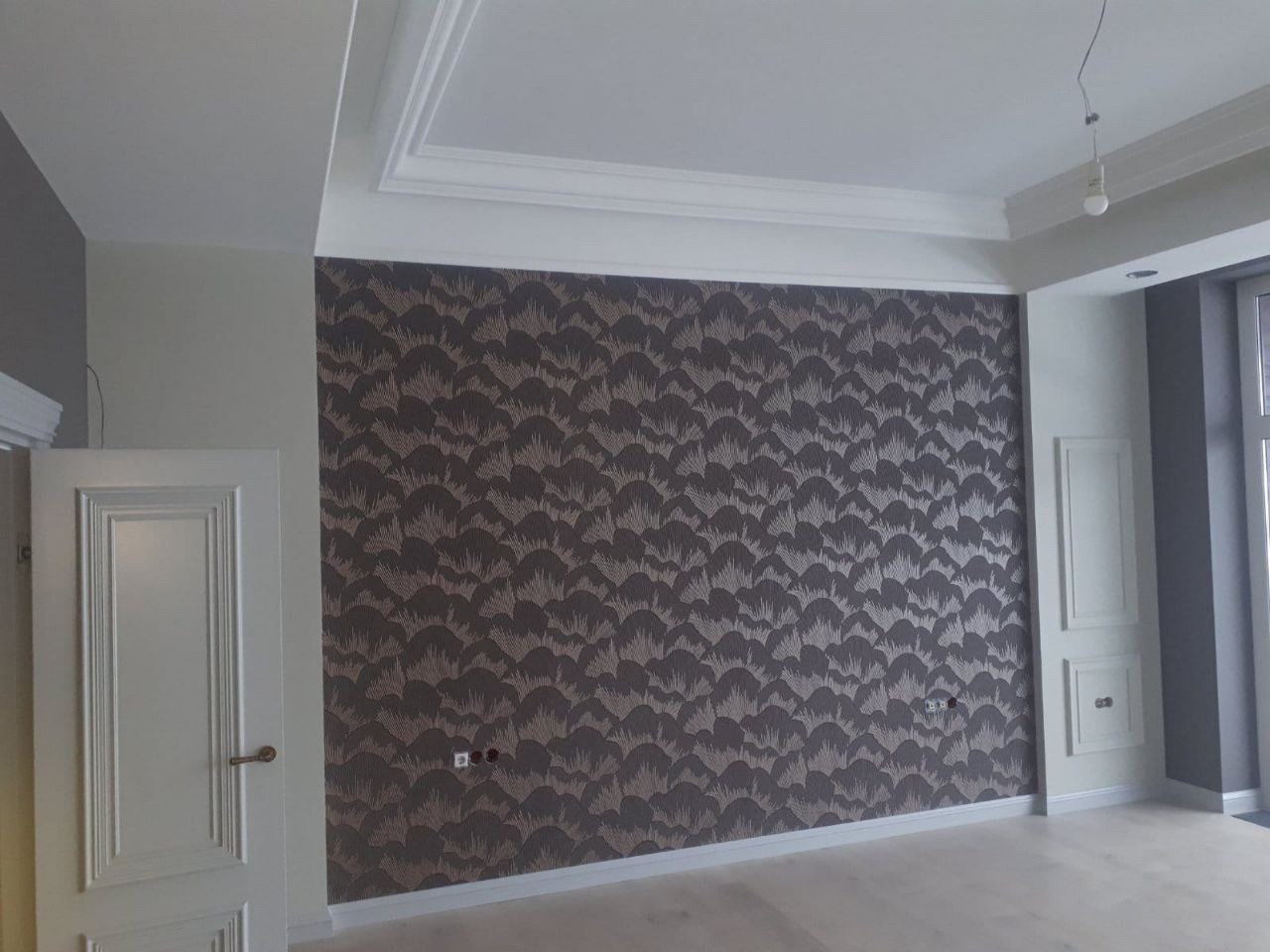Pokój po remoncie: ściana pokryta wzorzystą tapetą w odcieniach brązu i beżu, białe listwy przypodłogowe i sufitowe, białe drzwi z ozdobnymi panelami, gładka podłoga, widoczne gniazdka elektryczne.