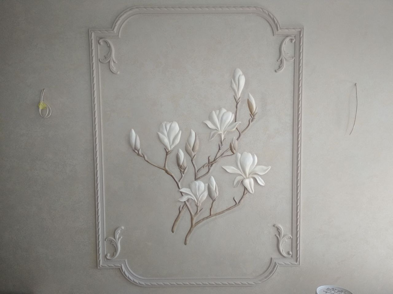 Dekoracyjna płaskorzeźba magnolii na gładkiej, beżowej ścianie w ozdobnej ramie ze sztukaterii.