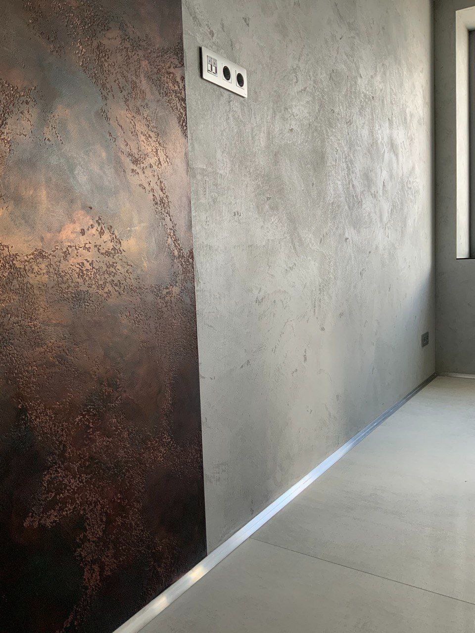 Ściana z dekoracyjną, miedzianą fakturą po lewej i szarą, gładką po prawej stronie, oddzielone aluminiową listwą przypodłogową, widoczne gniazdka elektryczne.