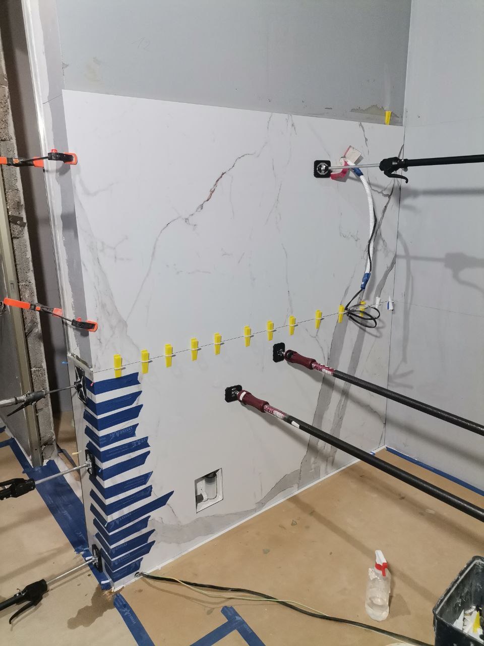 Montaż wielkoformatowych płyt imitujących marmur w łazience, użycie przyssawek i rozpórek do utrzymania płyty, widoczne poziomowanie z użyciem klipsów i pasków niebieskiej taśmy malarskiej...