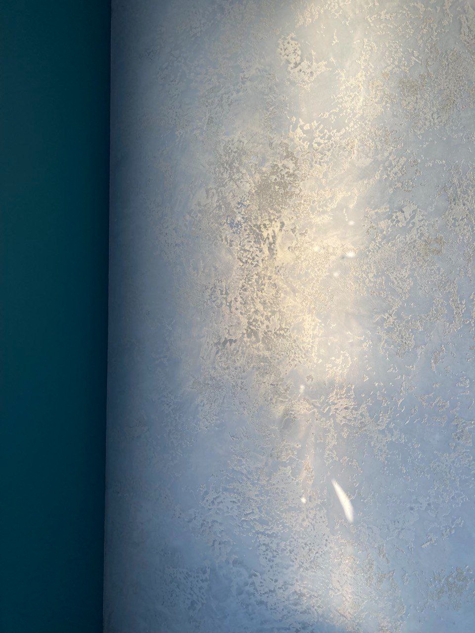 Dekoracyjna ściana z efektem perłowej poświaty i nieregularną fakturą, kontrastująca z gładką, ciemnoturkusową ścianą po lewej stronie. Widoczne refleksy światła podkreślają unikalny wzór.