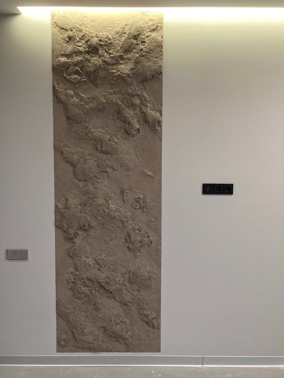 Dekoracyjna ściana z fakturą kamienia w pionowym pasie, podświetlona od góry, z szarymi włącznikami po bokach na gładkiej, białej ścianie.