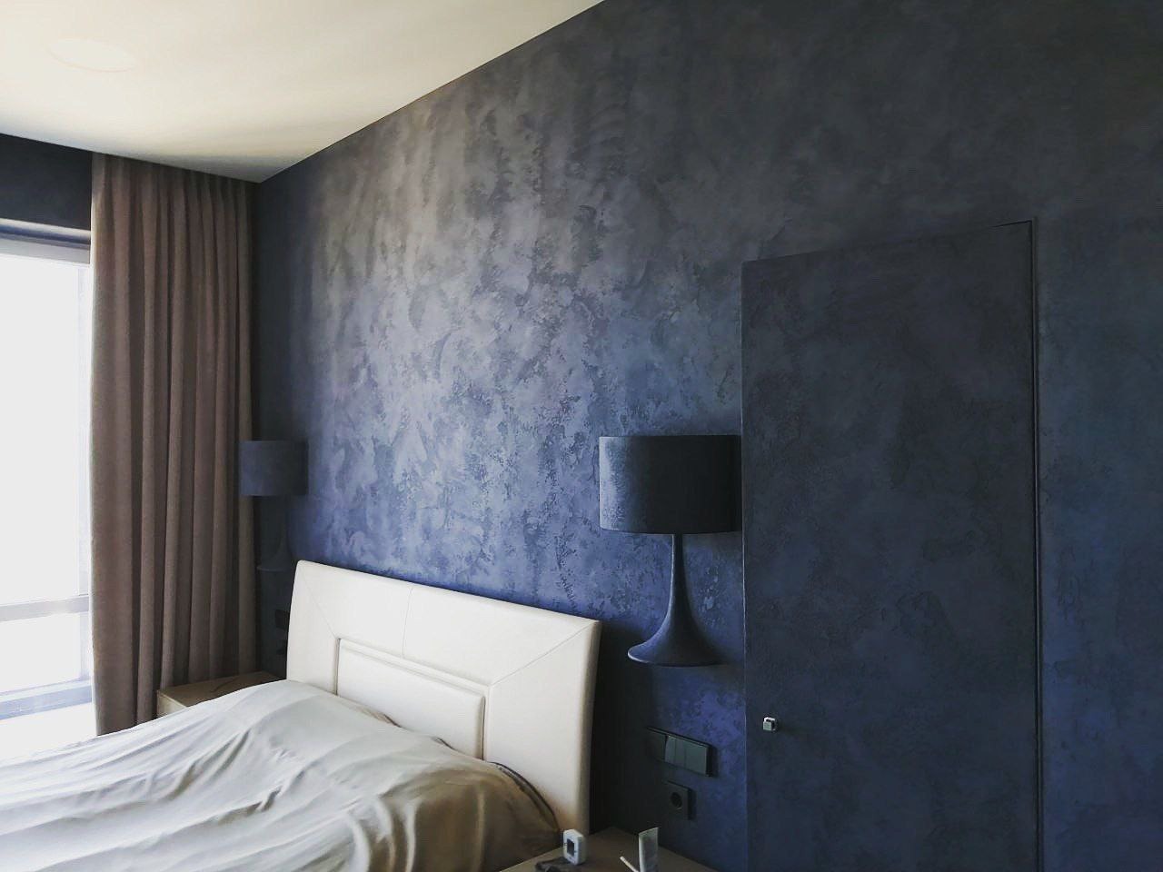 Sypialnia z ciemnoniebieską ścianą pokrytą dekoracyjną fakturą przypominającą chmury, kremowe wezgłowie łóżka i dopasowane zasłony w oknie.