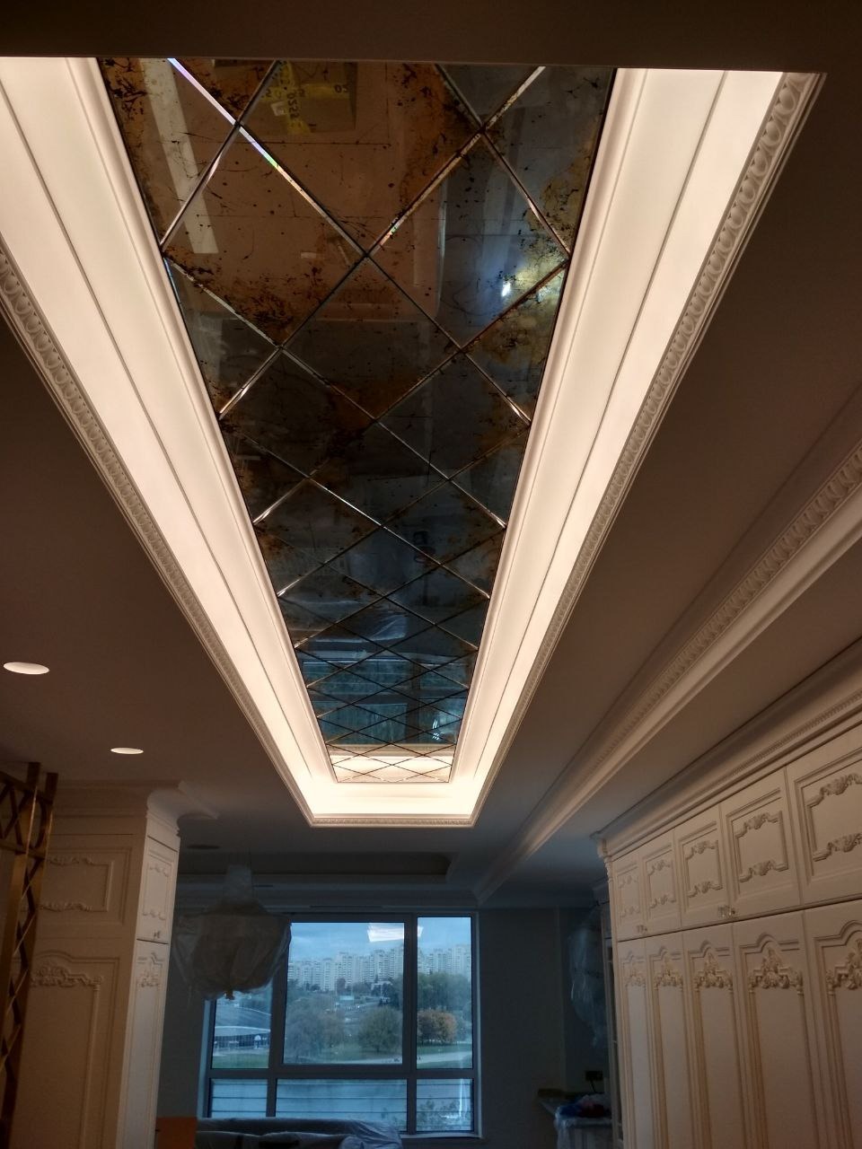 Dekoracyjny sufit z lustrzanymi kwadratami w romby, otoczony białym, oświetlonym sztukaterią, widok z dołu w salonie z dużym oknem.