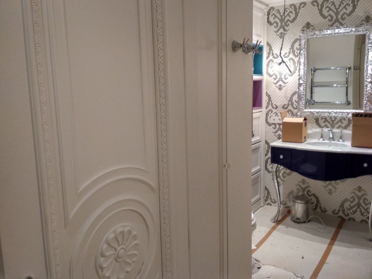 Fragment łazienki w trakcie wykańczania: widoczna biała, zdobiona szafa, umywalka z granatową szafką na srebrnych nóżkach, ozdobne lustro i mozaika na ścianie, kartony na blacie umywalki...