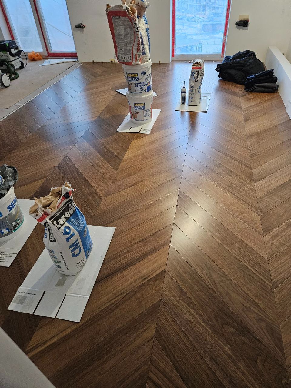 Układanie parkietu w jodełkę z widocznymi materiałami budowlanymi: worki z klejem i wiaderka z masą szpachlową, kartony ochronne na podłodze, okna w tle.