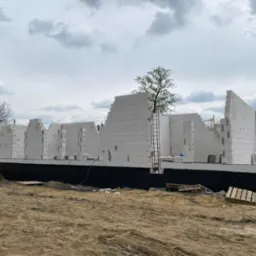 Wznoszone ściany domu z białych bloczków, widoczna czarna izolacja fundamentów, drabina oparta o ścianę, palety na piaszczystym podłożu, pochmurne niebo w tle.