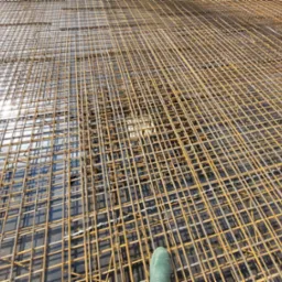 Siatka zbrojeniowa ułożona na szarym podłożu, widoczny fragment zielonego buta w prawym dolnym rogu.