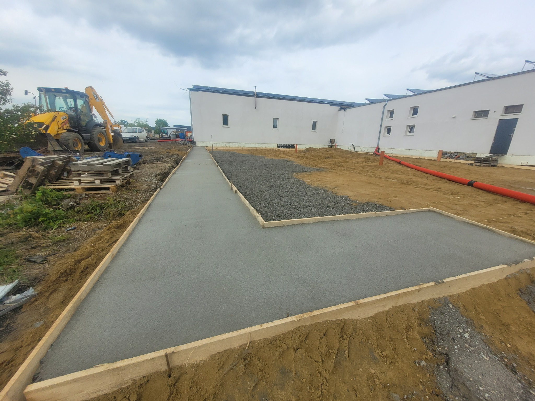 Wylany fundament pod budynek z widoczną deską szalunkową, częściowo zasypany kamieniem, z koparką w tle i budynkiem z panelami słonecznymi w tle.