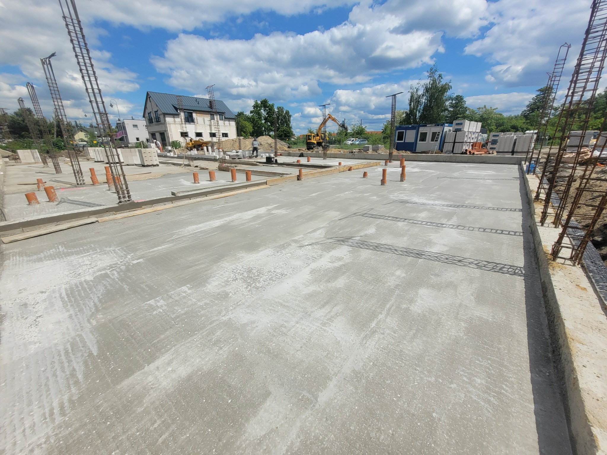 Wylana płyta fundamentowa z wystającymi rurami instalacyjnymi i zbrojeniami pionowymi. W tle dom w budowie i koparka. Słoneczny dzień.