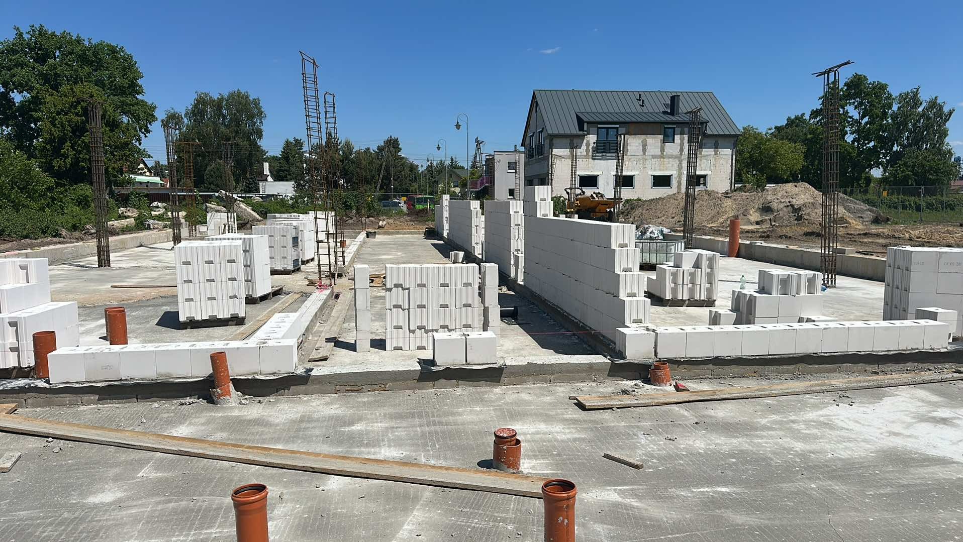 Budowa domu: fundamenty z betonu i bloczków Ytong, zbrojenie pionowe. W tle dom w stanie surowym, ziemia i zieleń. Słoneczny dzień, plac budowy w trakcie prac.