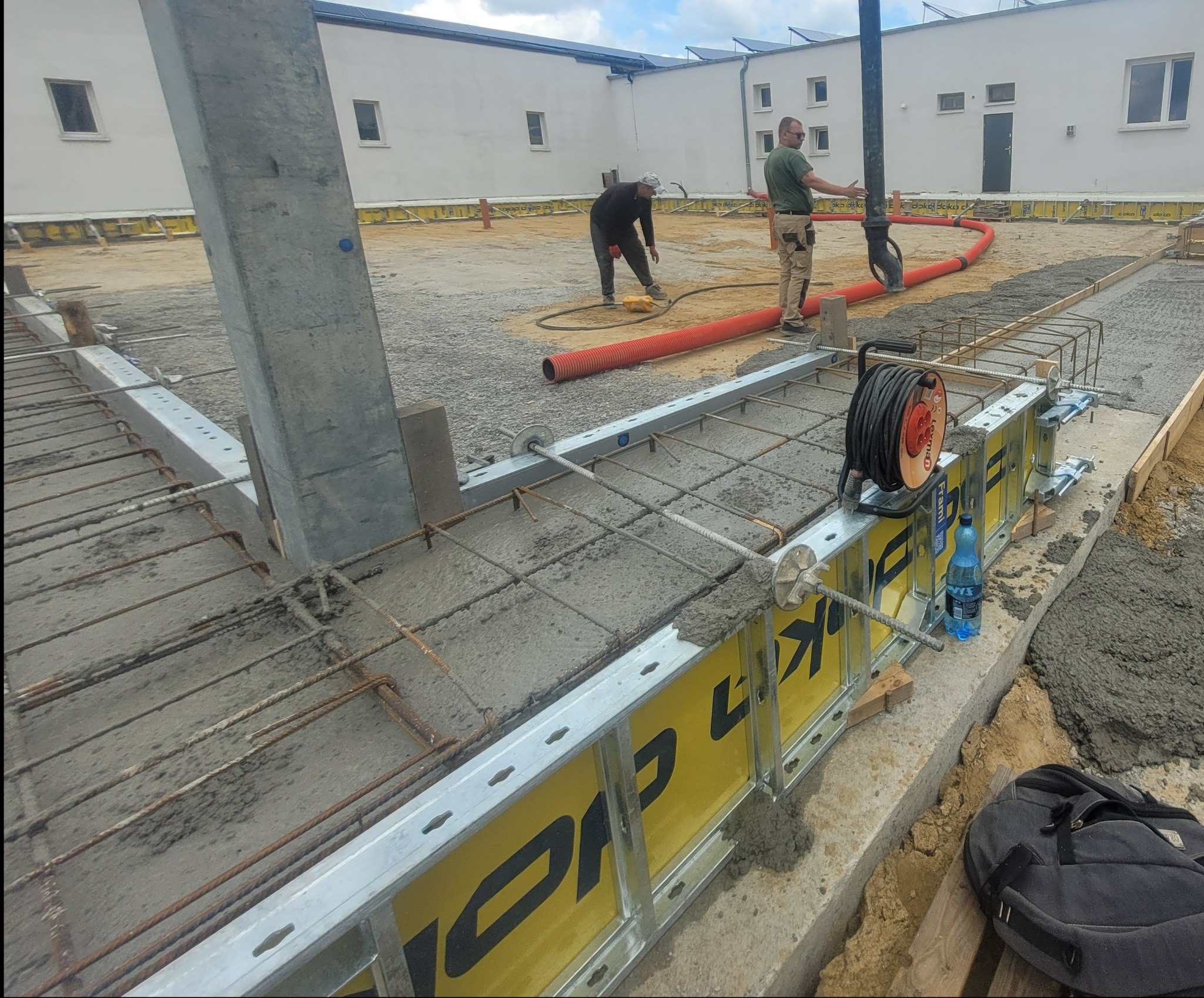 Wylewanie betonu na zbrojenie fundamentów budynku. Widoczne szalunki Doka, pracownicy i pompa do betonu. Etap budowy hali lub budynku żelbetowego.