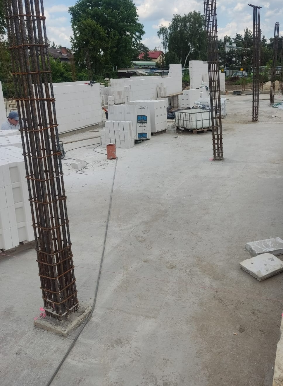 Wznoszenie ścian z białych bloczków i przygotowanie zbrojenia kolumn na płycie fundamentowej, widoczne materiały budowlane i częściowo zarys budynku w tle.