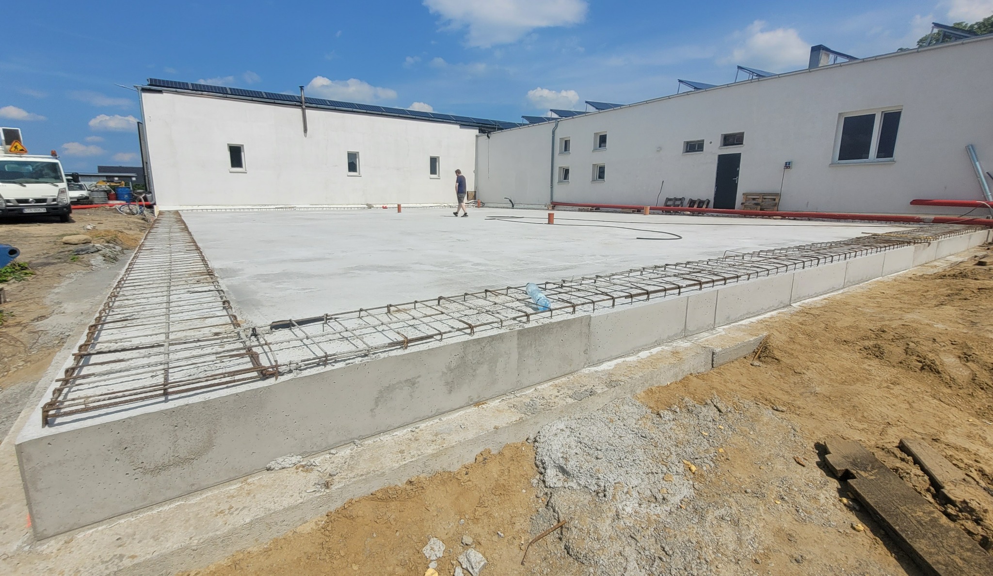 Wylana płyta fundamentowa budynku z widocznym zbrojeniem obwodowym i fragmentem konstrukcji budynku w tle, w słoneczny dzień.