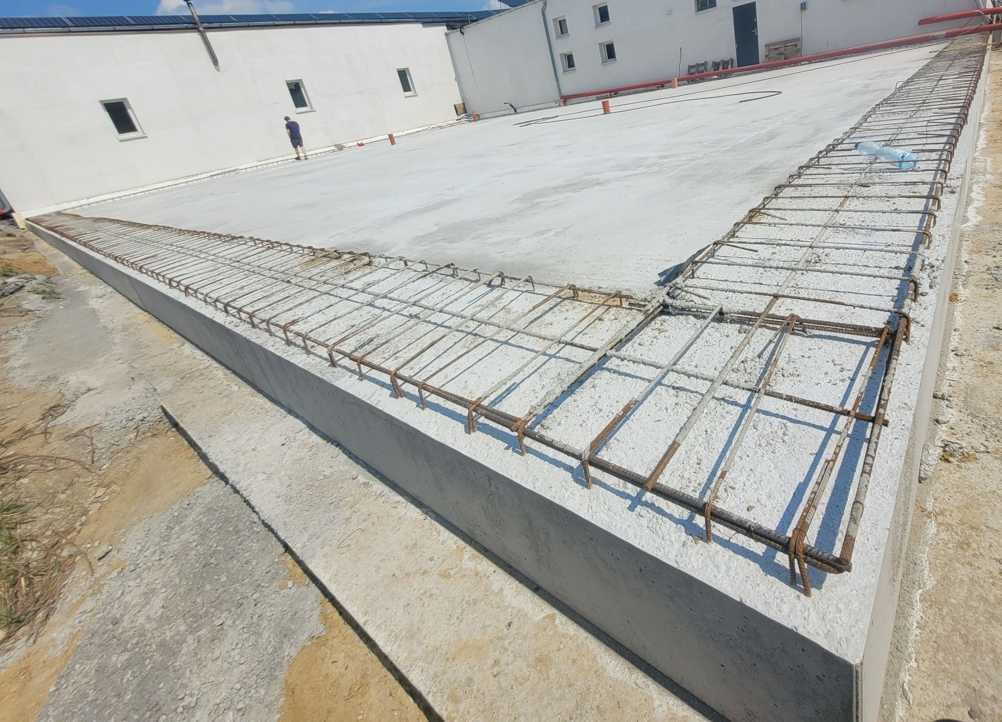 Zbrojenie obwodowe na betonowym fundamencie budynku, widoczne pręty zbrojeniowe i fragment budynku w tle. Na fundamencie leży pusta butelka PET.