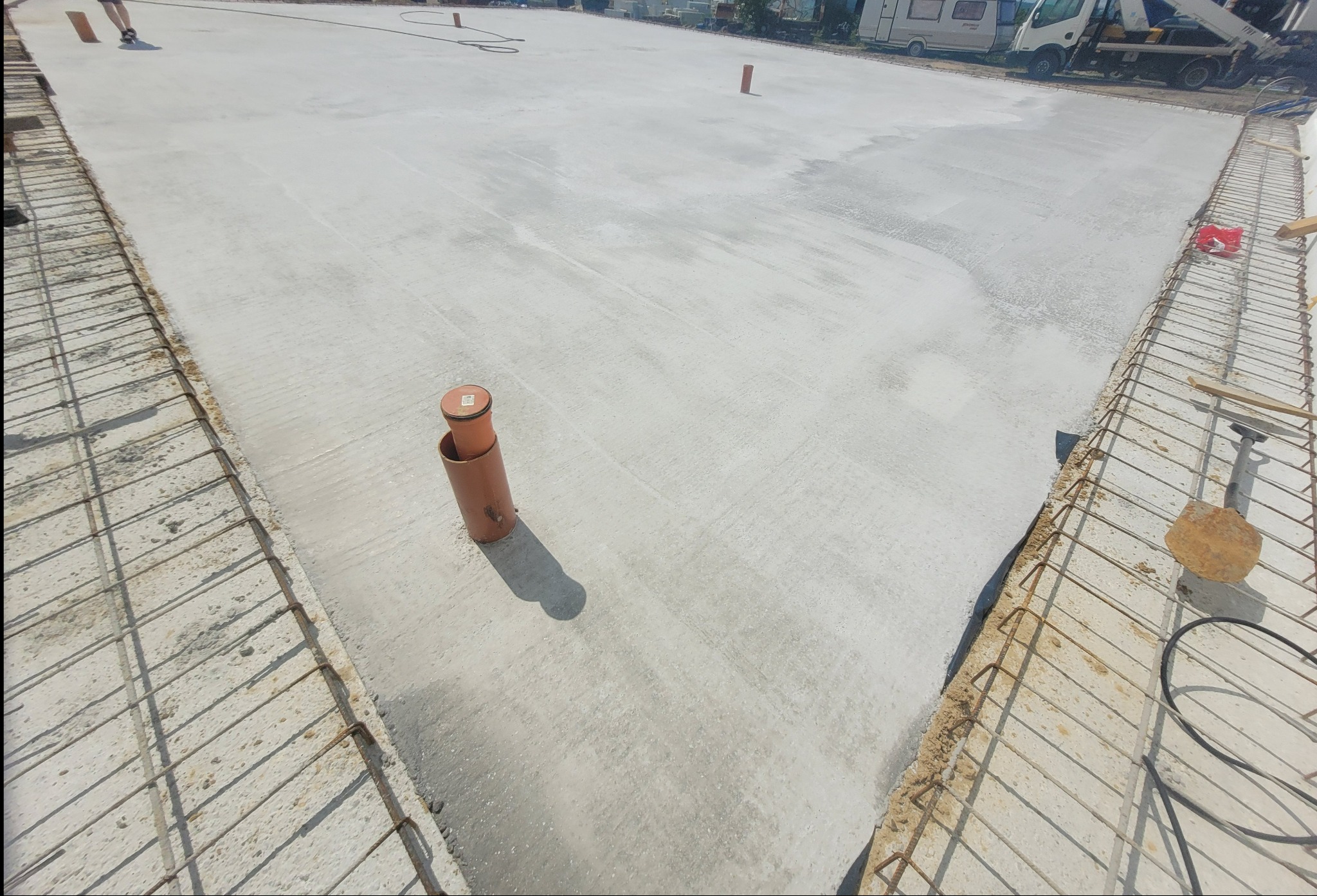 Świeżo wylany fundament z wystającymi rurami kanalizacyjnymi i zbrojeniami po bokach. Widoczne ślady wilgoci na powierzchni betonu.