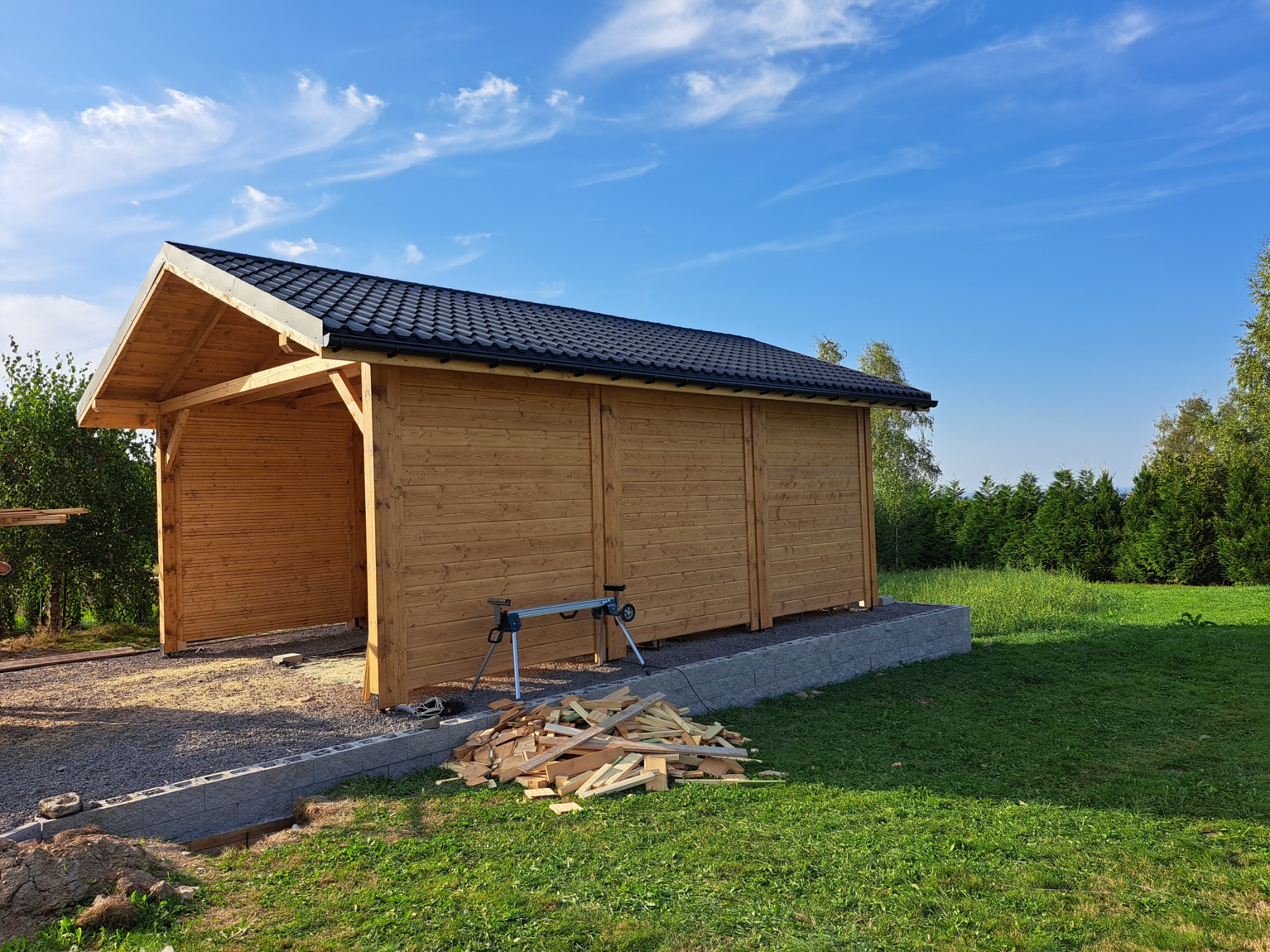 Nowo wybudowana, drewniana konstrukcja z czarnym dachem na tle błękitnego nieba z chmurami, widoczne narzędzia i pozostałości drewna, sugerujące trwające prace wykończeniowe.