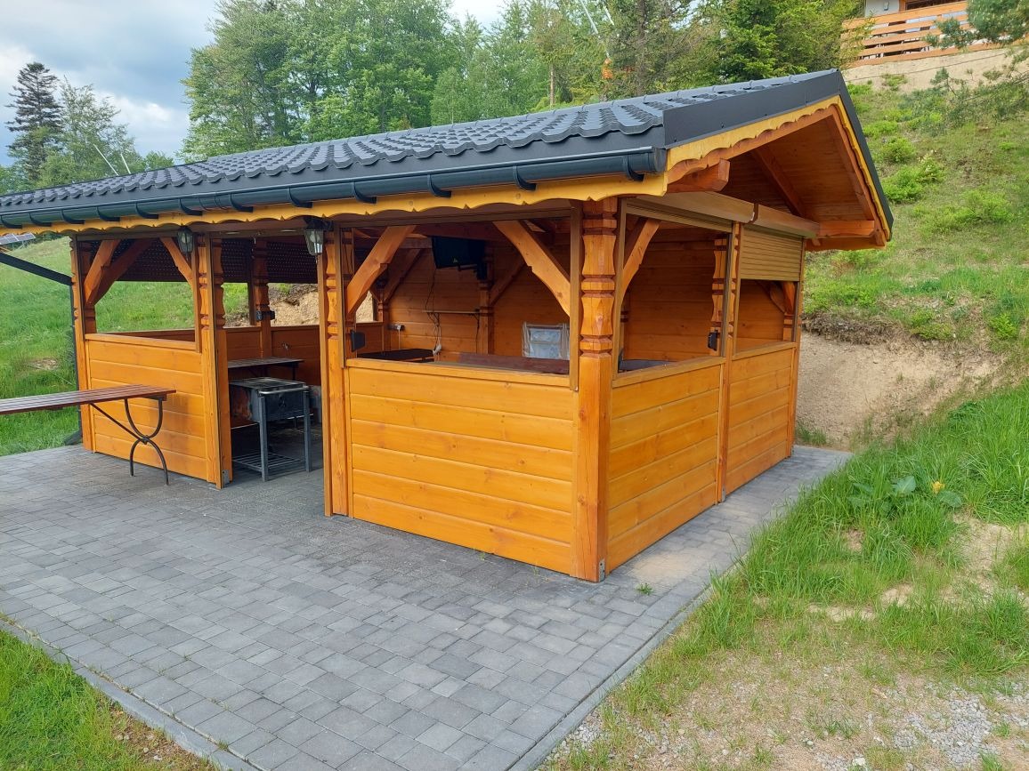 Drewniana altana grillowa z czarnym dachem w Szczyrku, z wyposażeniem do grillowania i stołem na zewnątrz, na wybrukowanym terenie.