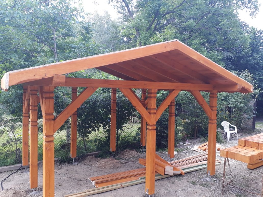 Konstrukcja z drewna o pomarańczowym odcieniu, prawdopodobnie w trakcie budowy, widoczne podpory, belki i zadaszenie, w tle zieleń drzew i krzewów, oraz białe krzesło.